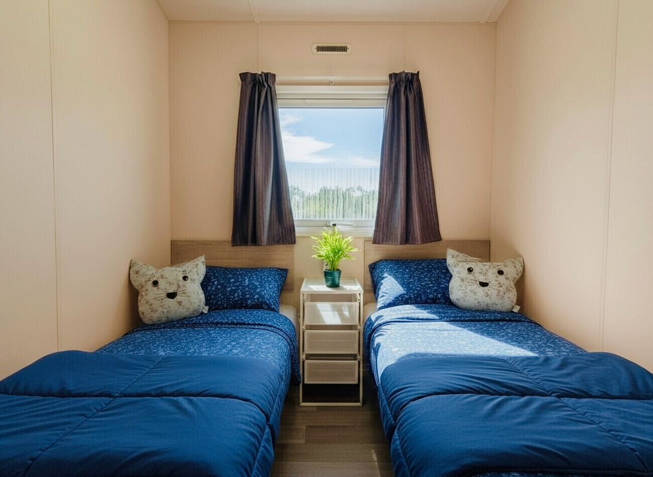 Una habitación con dos camas, cada una con ropa de cama azul a juego y almohadas con motivos de gatos. En el centro, hay una pequeña mesa con una planta.