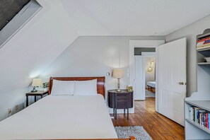 Ein Schlafzimmer mit einem großen Bett, einem Nachttisch mit Lampe, einem kleinen Beistelltisch, einem Bücherregal und einer Tür zu einem weiteren Raum.