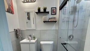 A bathroom with a toilet, sink, and shower. There are shelves with decorative items and a mirror above the sink.