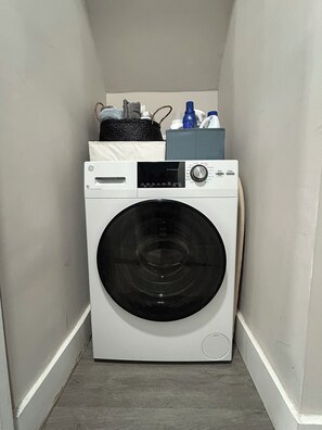 A compact, white front-loading washing machine in a narrow laundry room with a small storage cabinet above it. - Peaceful Garden Apartment on Quiet Brooklyn Street (BROOKLYN)