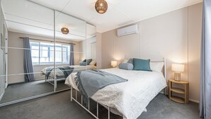 Une chambre moderne avec un grand miroir, un lit recouvert d’une couette blanche, une lampe de chevet et une petite table de nuit en bois.