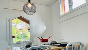 Un coin repas avec une table en bois, des chaises blanches et une vue sur les maisons par la fenêtre.