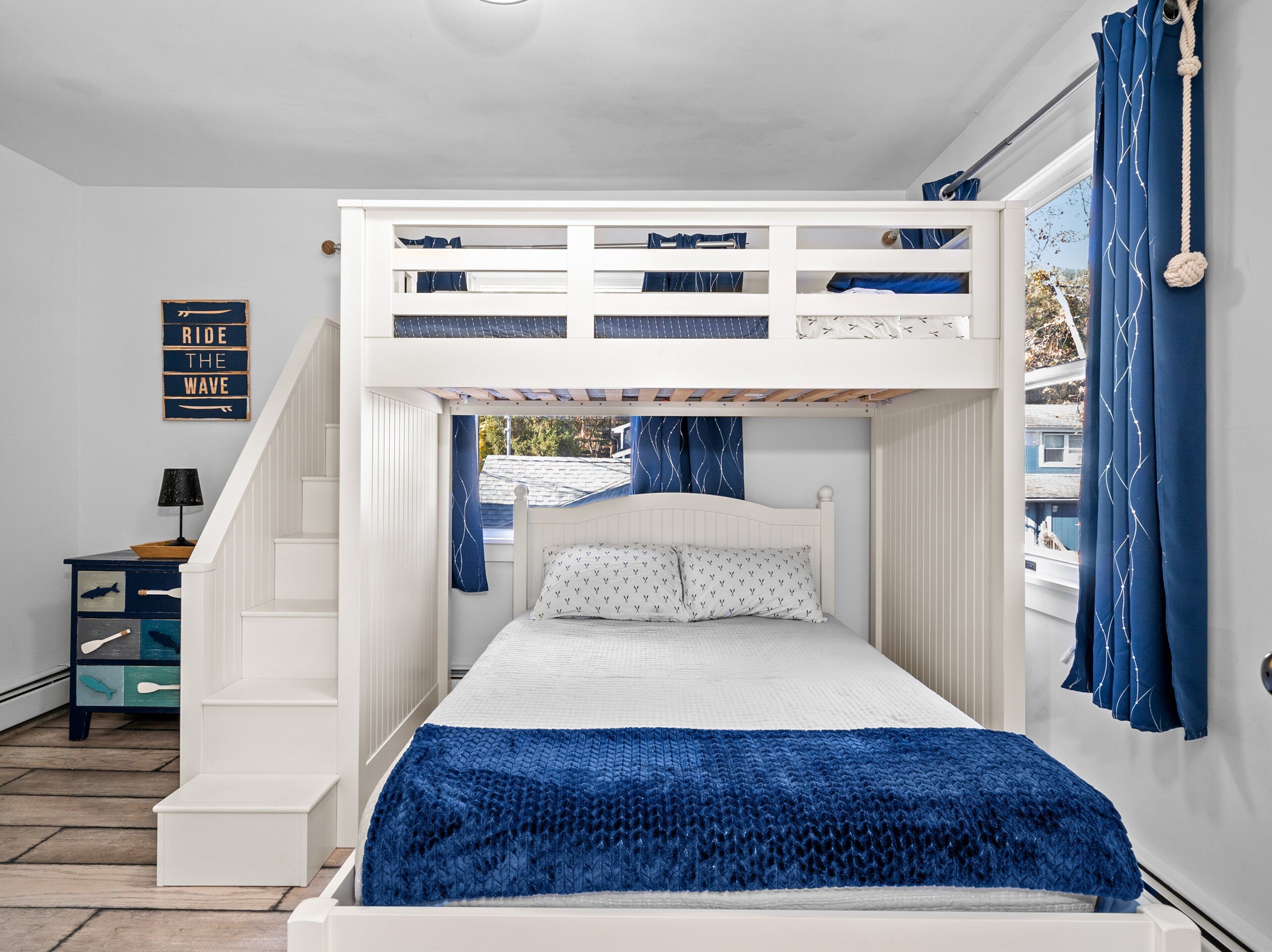 A bunk bed with a white frame and blue bedding, a small table with a lamp, and a 'Ride the Wave' sign on the wall.