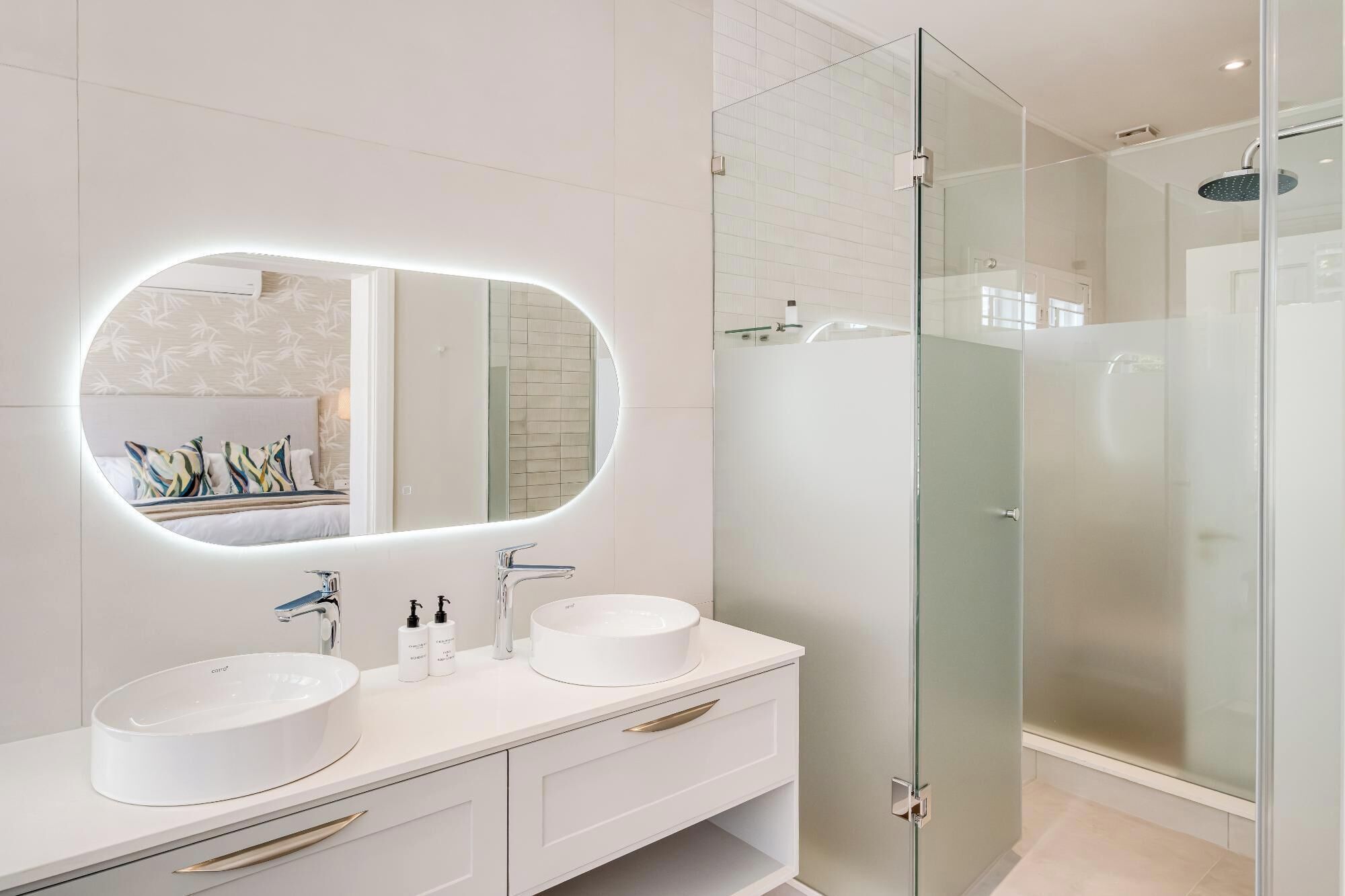 A modern bathroom with a large oval mirror, two sinks, and a glass-enclosed shower.