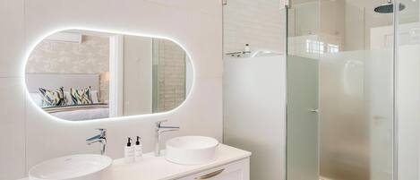 A modern bathroom with a large oval mirror, two sinks, and a glass-enclosed shower.