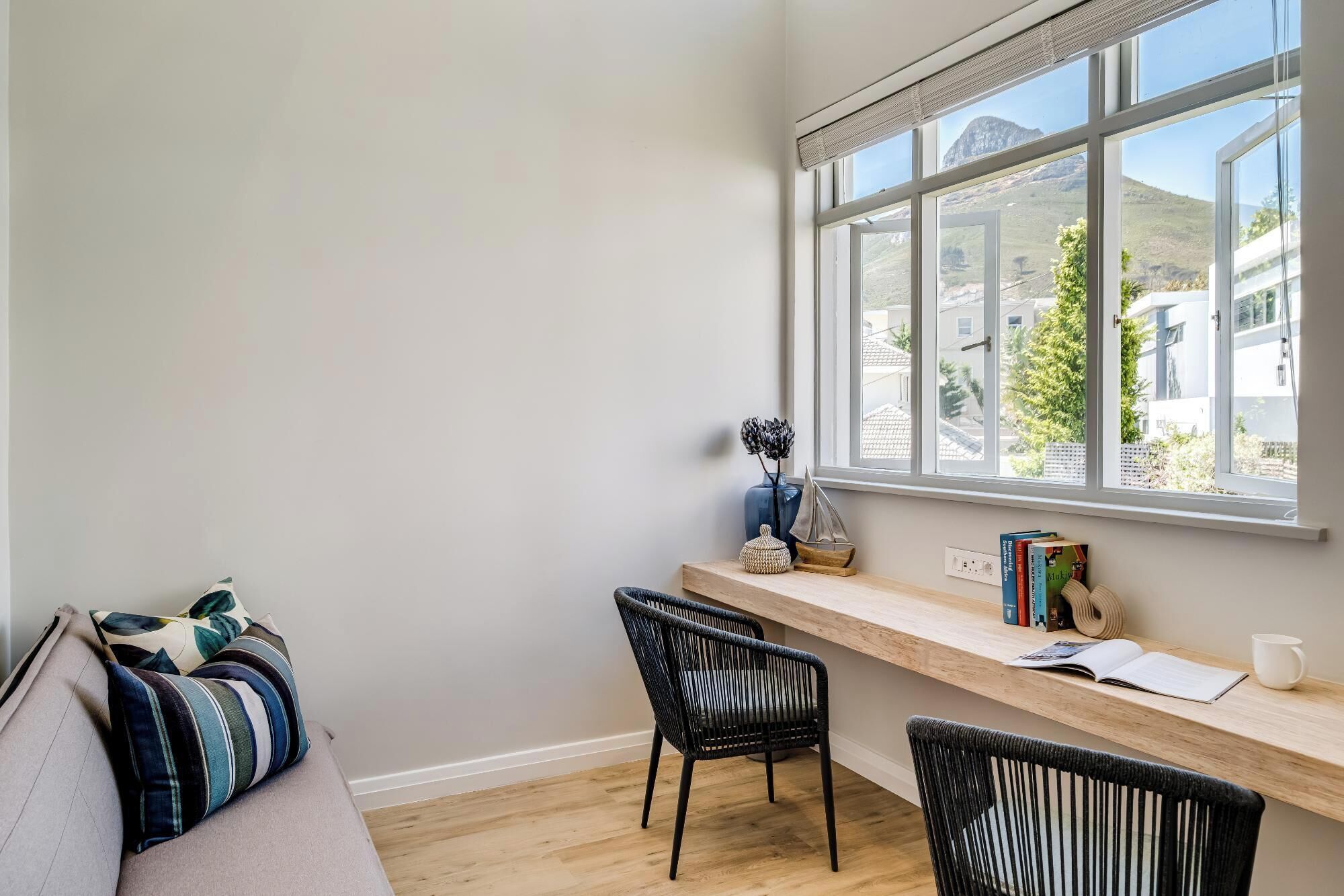 A cozy reading nook with a wooden desk, two black chairs, a cushioned bench, and a window with a view of houses and a mountain.