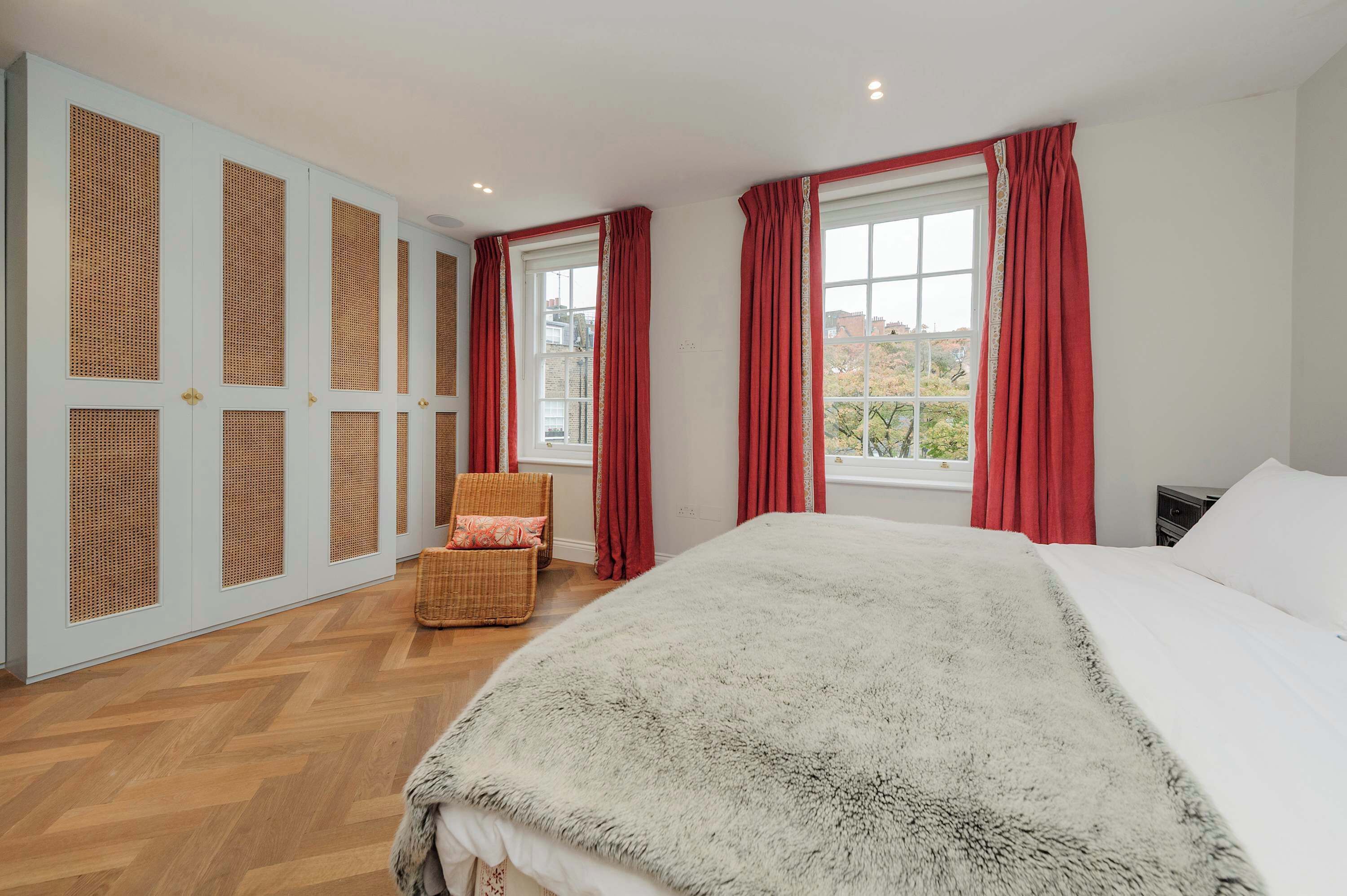 Une chambre avec un grand lit, une chaise en osier, une fenêtre avec des rideaux rouges et une vue sur l’extérieur.