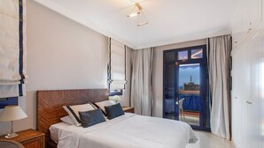 Ein Hotelzimmer mit einem großen Bett, einem Holzkopfende, einem Nachttisch mit Lampe, Blick durch eine Glastür nach draußen und einem Balkon.