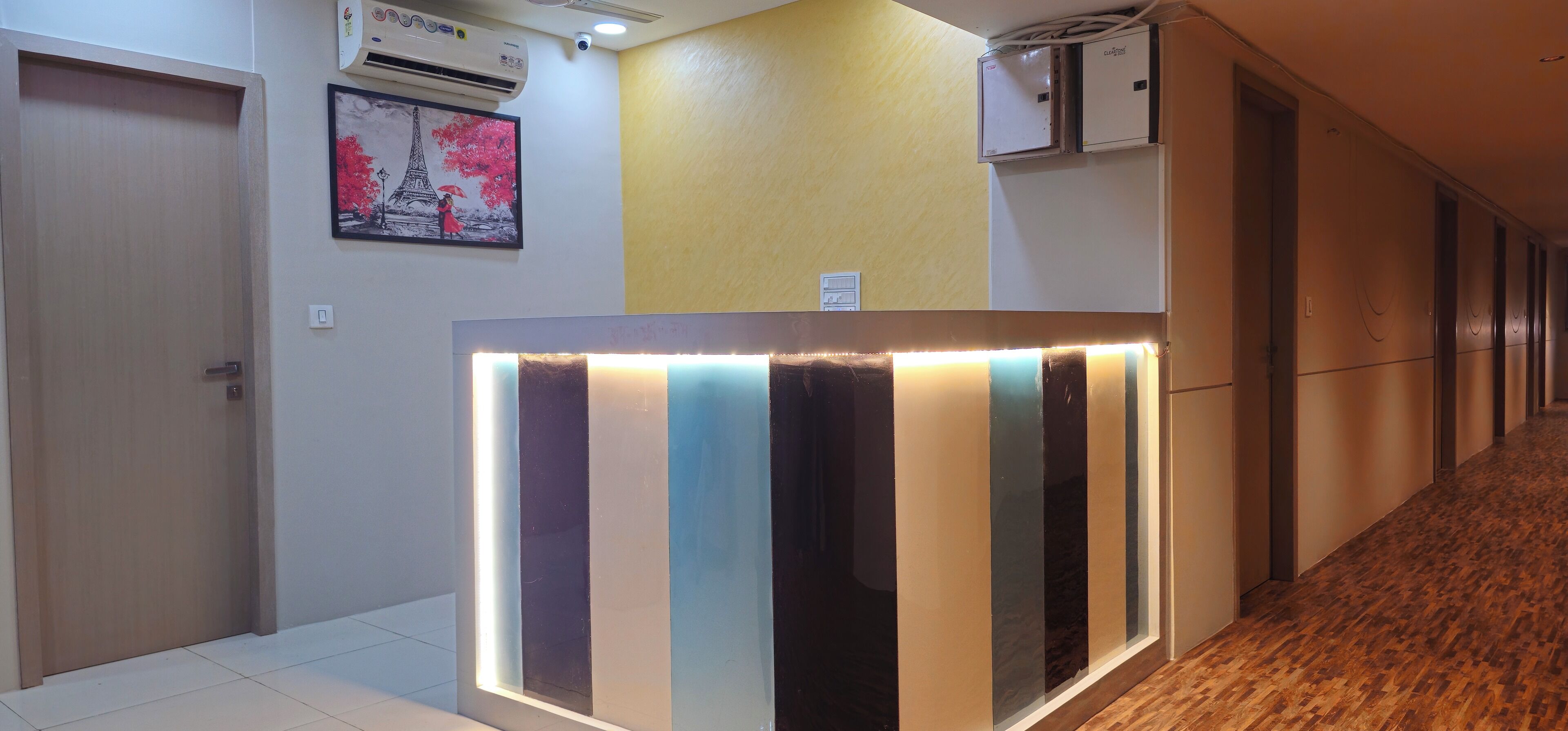 A hotel reception area with a modern design, featuring a reception desk with illuminated panels, a framed picture of the Eiffel Tower, and a yellow accent wall.