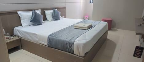 A neatly made bed with pillows, a bedside table with a glass jar and a tray, and a pink stool.