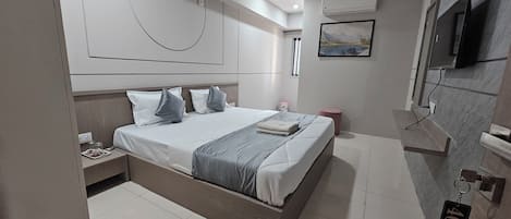 A modern hotel room with a large bed, a bedside table with a lamp, a mounted TV, and a painting on the wall.