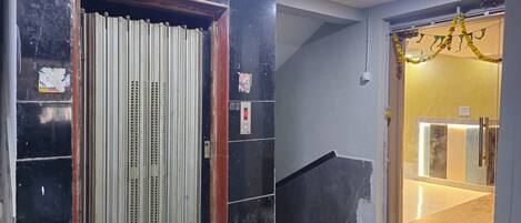 Ein Aufzug mit einer Metalltür, einer Bedienungsanlage und einer Treppe, die zu einem Raum mit einer Leuchte und einer Tür mit einem goldenen Girlande führt.