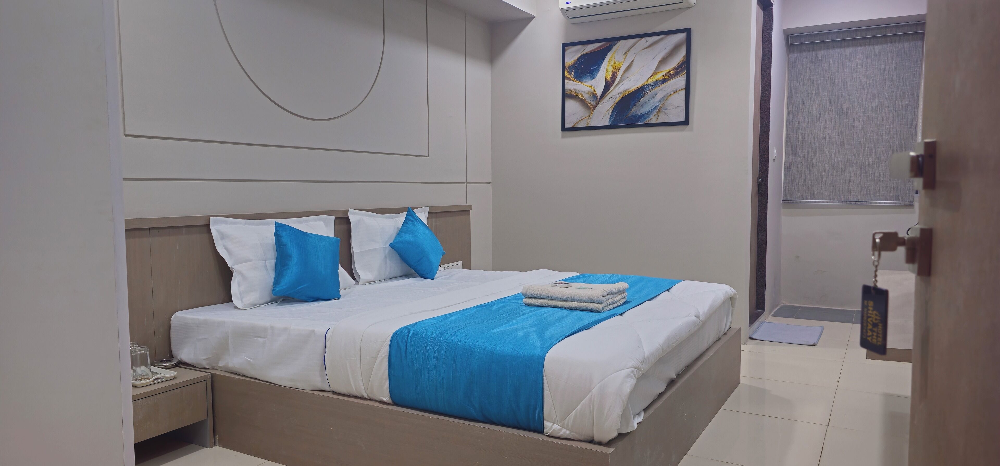A neatly made bed with white and blue bedding, a bedside table with a glass and a plate, and a wall-mounted air conditioner.