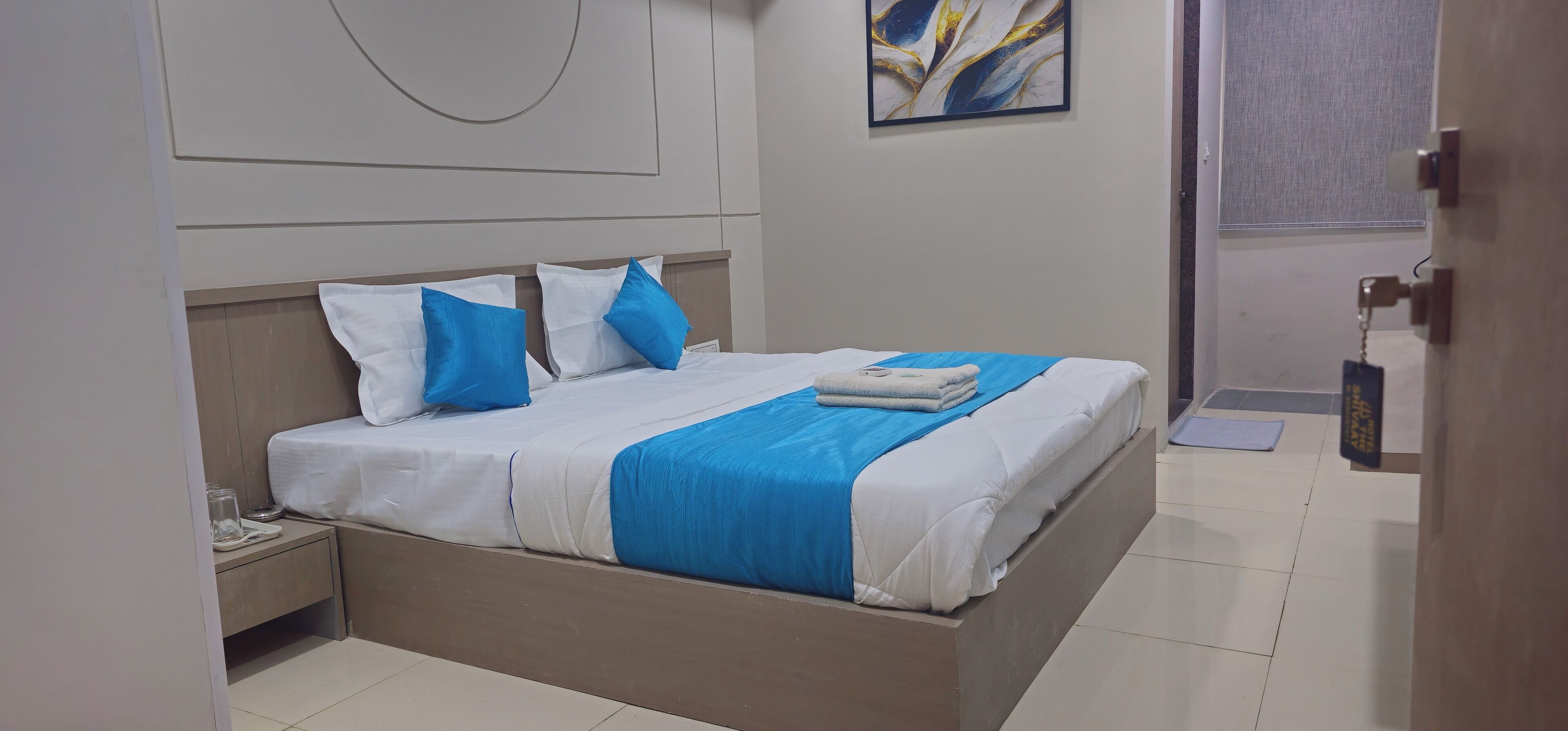 Een net opgemaakt bed met wit en blauw beddengoed, een nachtkastje met een glas, en een wanddecoratie.