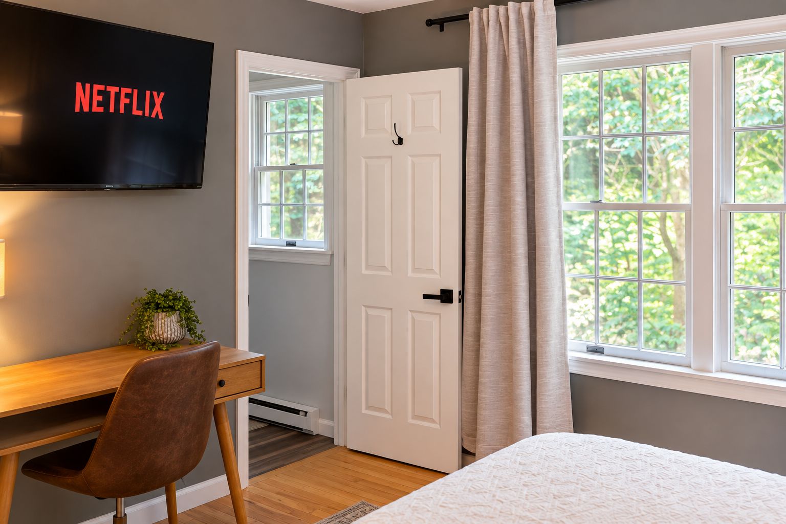 Une chambre à coucher comprenant un lit, un bureau avec une chaise, un téléviseur fixé au mur, une porte et une fenêtre avec des rideaux.