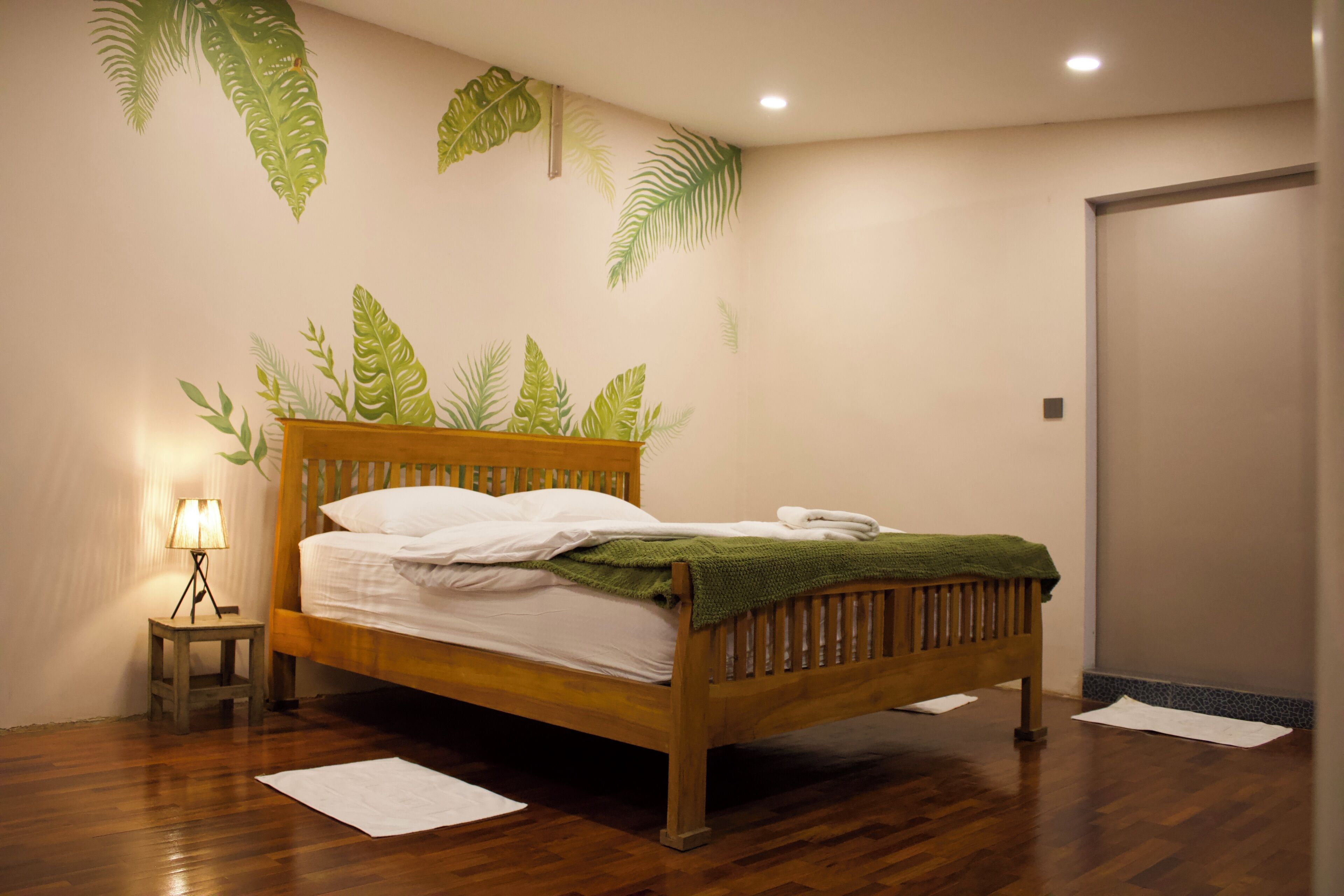 A neatly made bed with a wooden headboard, a small bedside table with a lamp, and a wall decorated with leaf patterns.