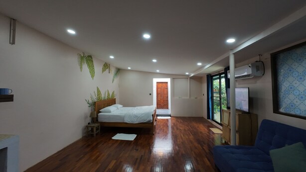 A bedroom with a wooden floor, a bed, a blue sofa, a TV, and a window with a view of greenery. - Oasis studio - Great location (Vientiane)