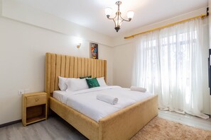 A neatly made bed with a wooden headboard, a small bedside table, and a large window with sheer curtains. - Luxurious Nairobi Apartment With City and Garden Views (Nairobi)