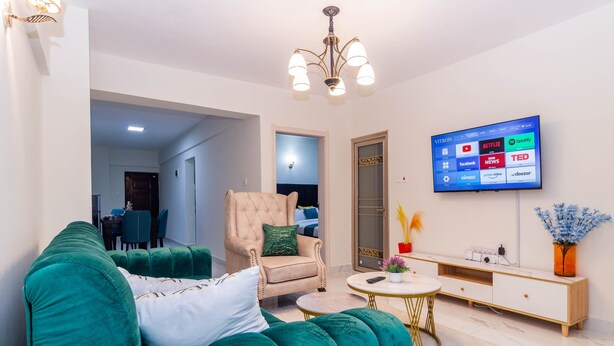 Ein Wohnzimmer mit einem grünen Sofa, einem beigefarbenen Sessel, einem kleinen runden Couchtisch und einem Fernseher, der Streaming-Dienste anzeigt.