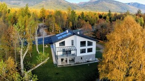 Une maison moderne à deux étages, dotée d’un toit plat, de grandes fenêtres et de panneaux solaires, entourée d’une forêt aux couleurs automnales et avec des montagnes en arrière-plan.