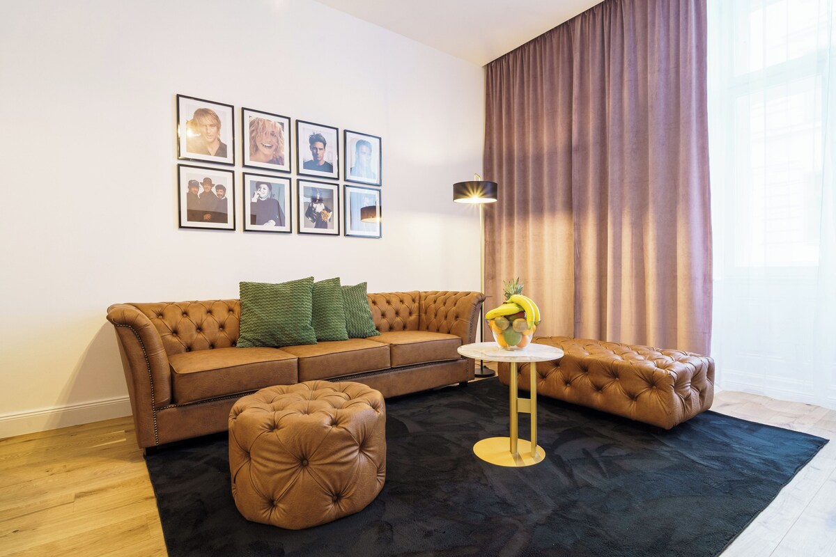 A living room with a brown sofa, a round coffee table, and a tufted ottoman. There are framed pictures on the wall and a lamp on the table.