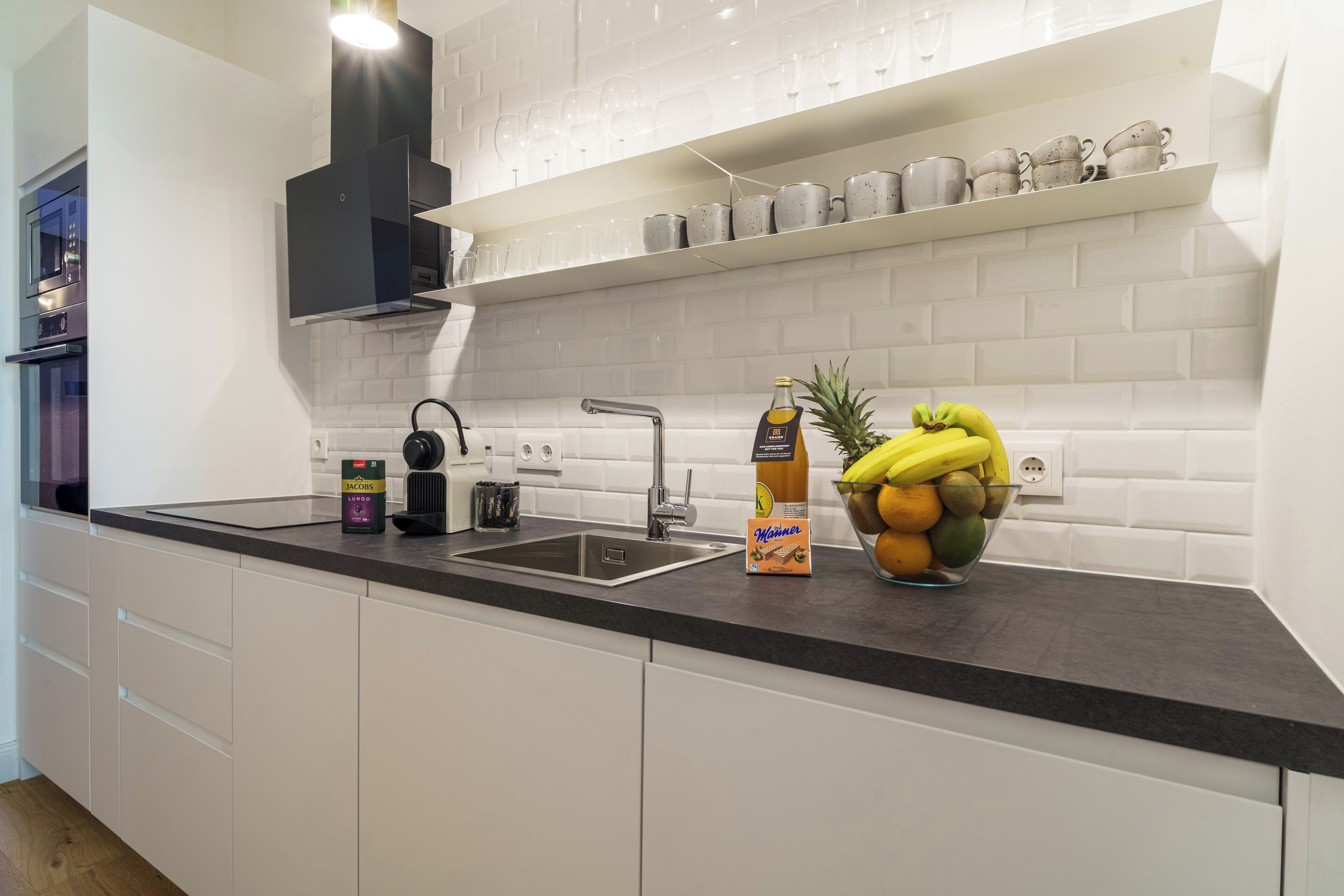 Une cuisine moderne avec des meubles blancs, un plan de travail noir, un évier en acier inoxydable et un panier à fruits.