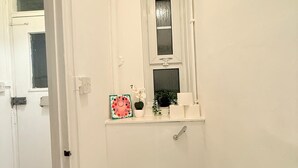 Une petite salle de bain blanche, avec des toilettes, une fenêtre et une étagère sur laquelle sont posés un pot de fleurs et un tableau encadré.