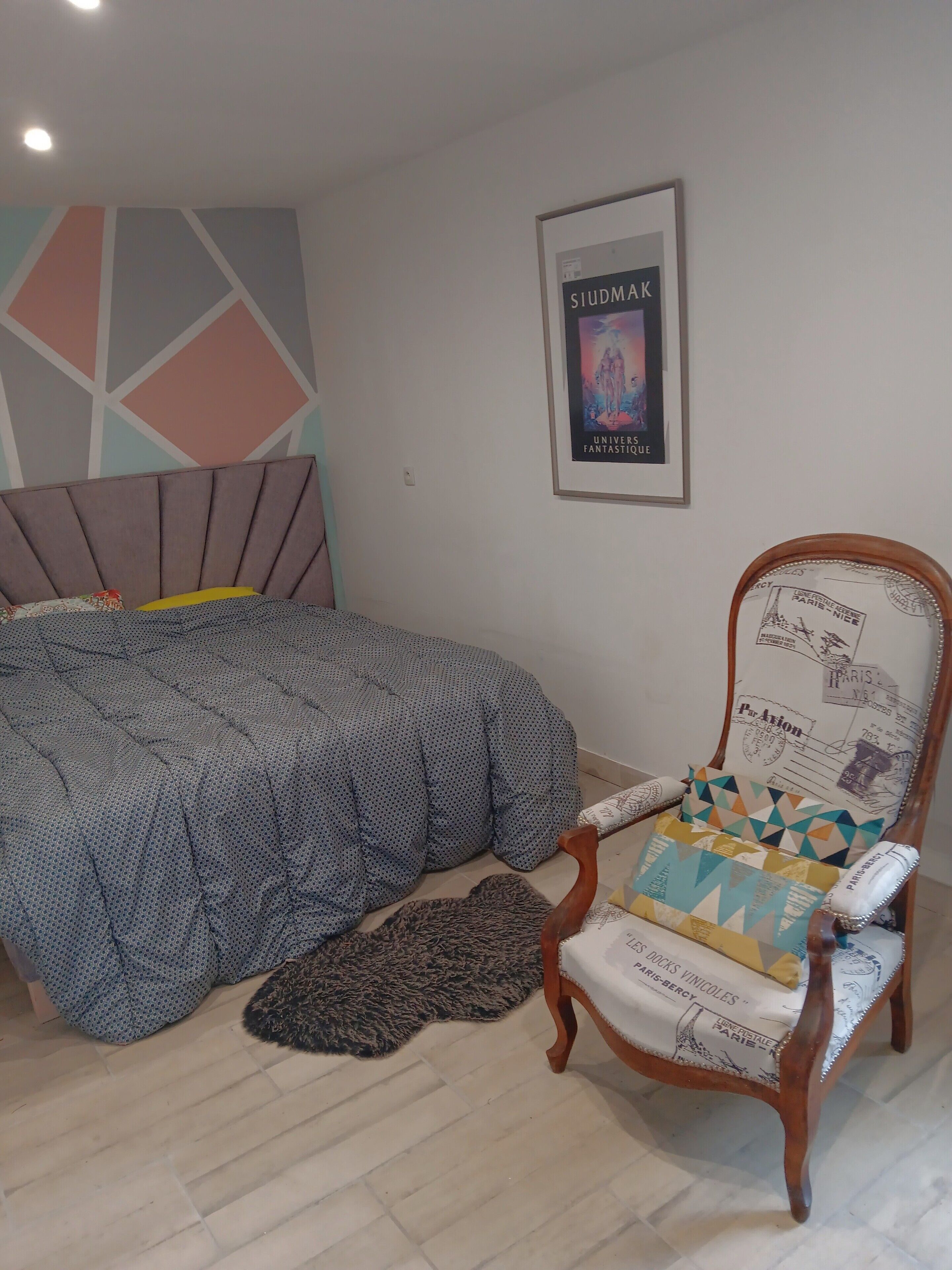 Ein Schlafzimmer mit einem Bett, einem Sessel mit gemustertem Kissen, einem Teppich und einem Bild an der Wand.