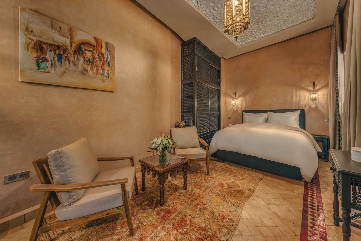 A hotel room with a large bed, two armchairs, a small table with a vase, a patterned rug, and a painting on the wall.