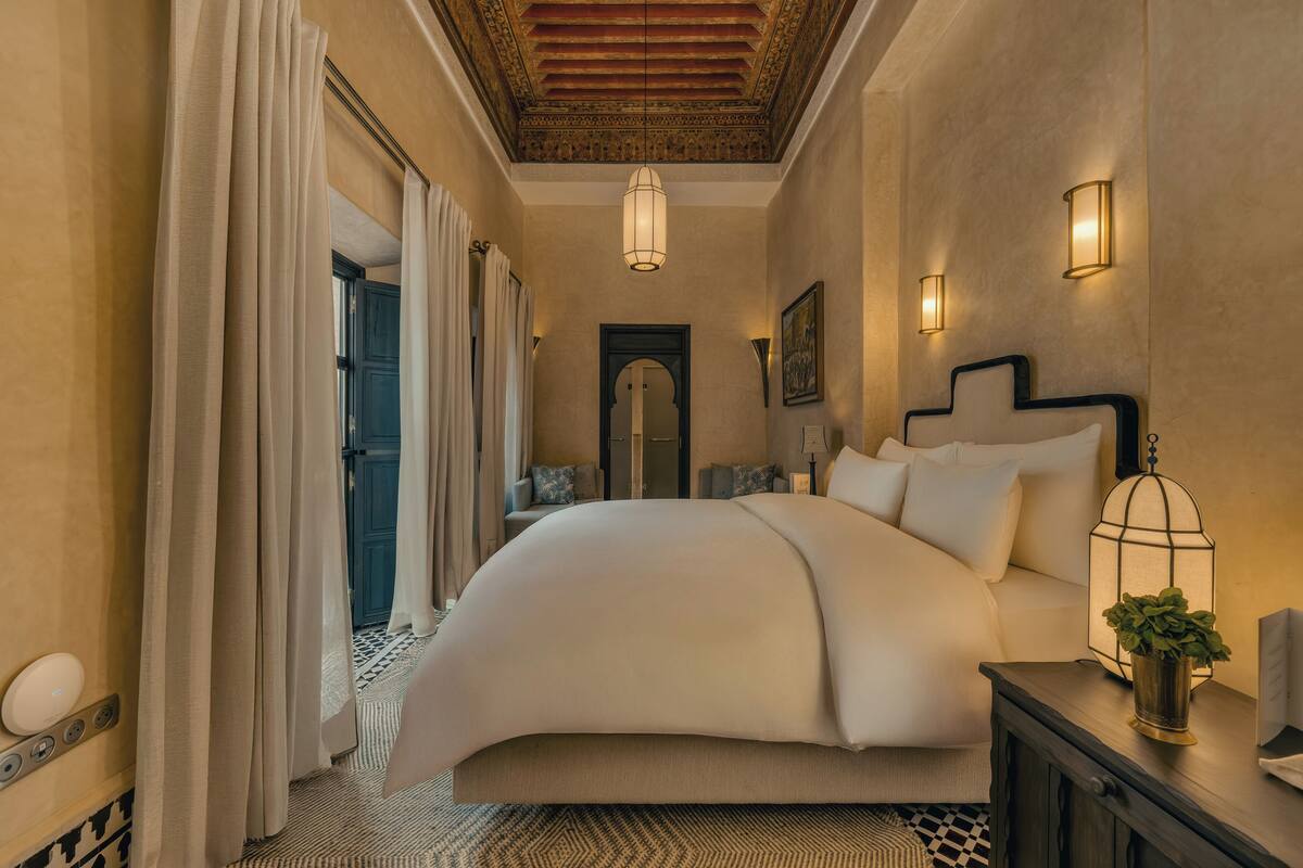 A spacious bedroom with a large bed, a nightstand with a lamp, and a view of the hallway.