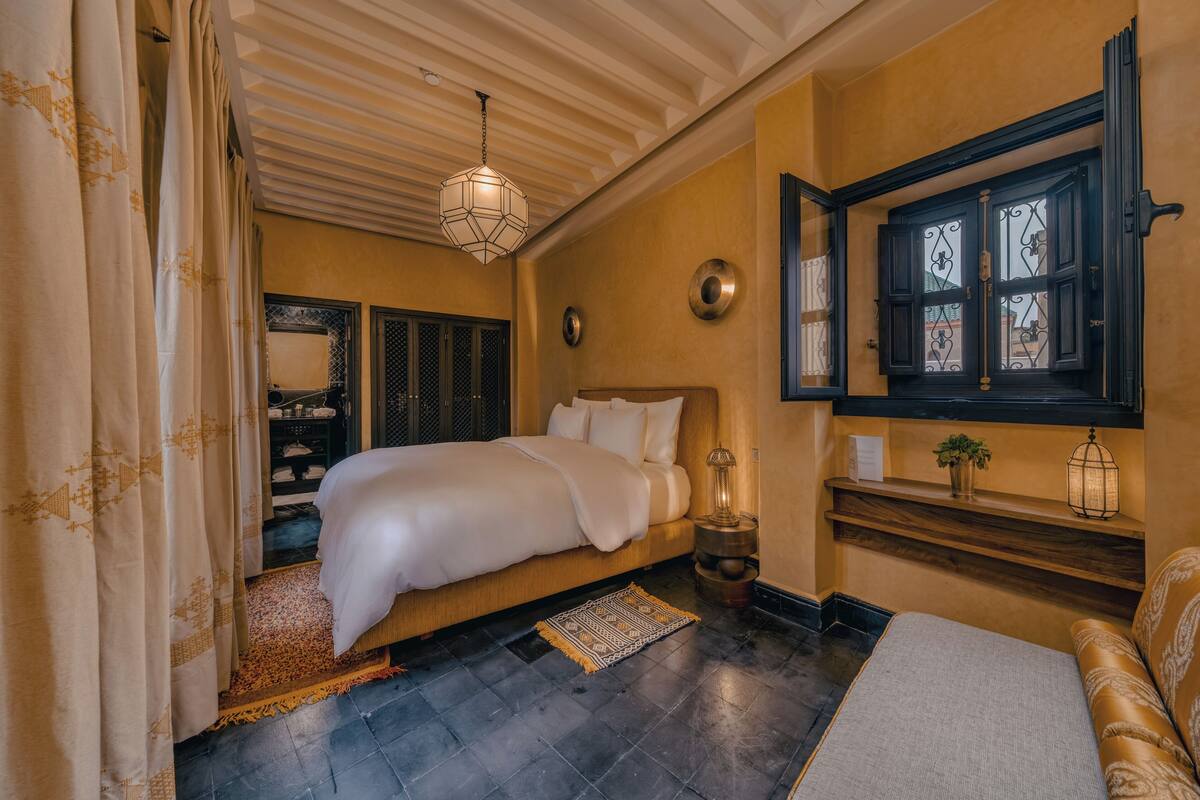 A bedroom with a bed, a nightstand, a lamp, a window with decorative shutters, and a small shelf with a plant.