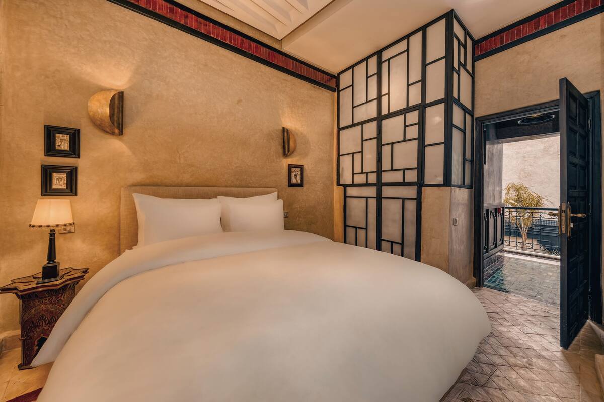 A large bed with white linens, a nightstand with a lamp, and a wall-mounted light fixture.