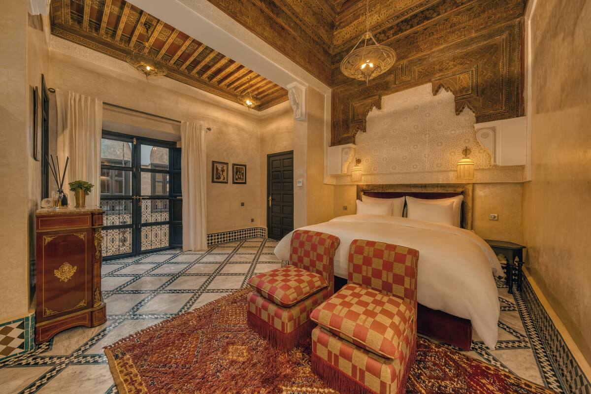 A bedroom with a large bed, checkered ottomans, a wooden ceiling, and patterned floor tiles.