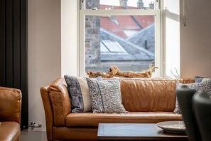 A brown leather sofa with patterned cushions, a wooden coffee table, and a view of a building through a window. - Anderson's Neuk | Anstruther (Anstruther)