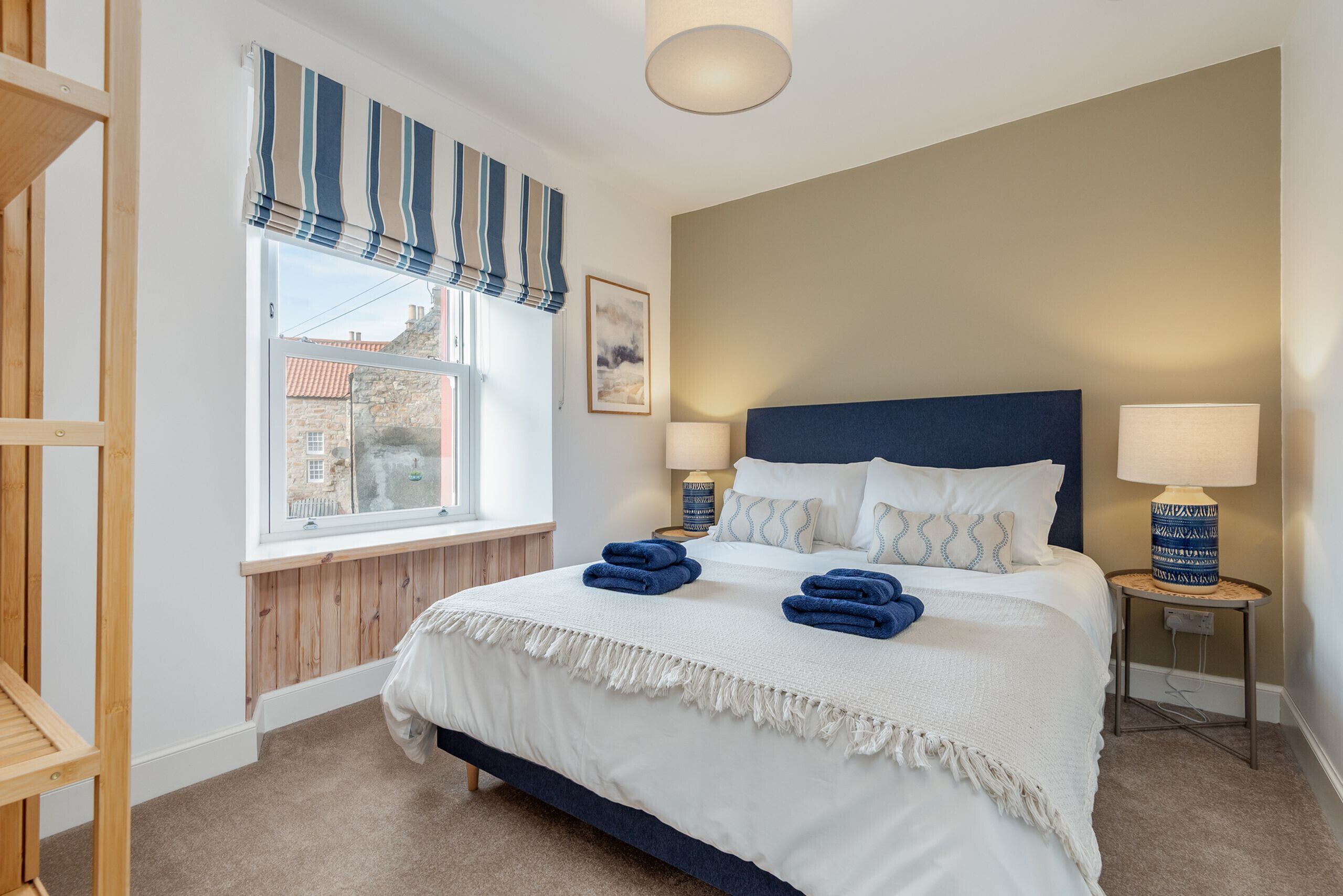 A bedroom with a bed, two bedside tables, a window with curtains, and a view of buildings outside.