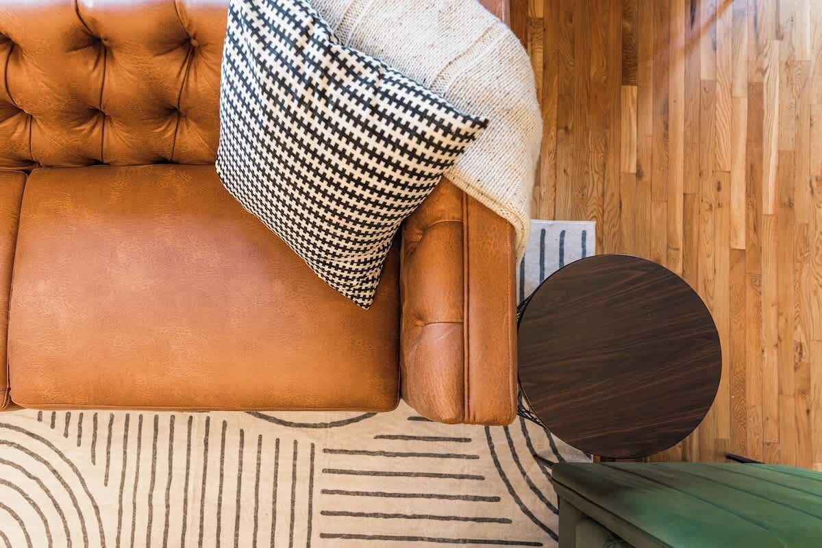 A brown leather sofa with a houndstooth pillow, a round wooden side table, and a patterned rug.