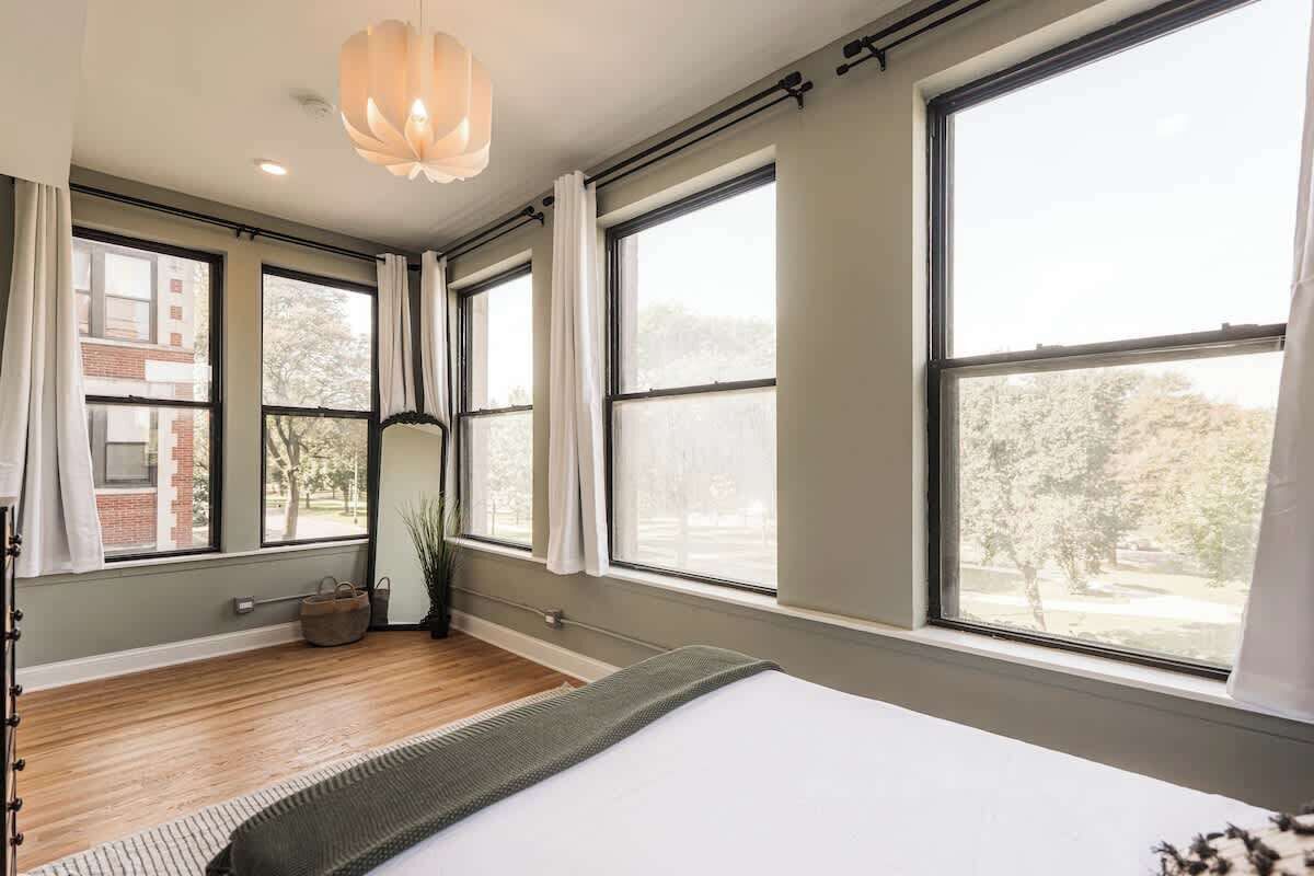 Une chambre avec de grandes fenêtres, un lit, un luminaire suspendu et une vue sur des arbres et un bâtiment à l’extérieur.