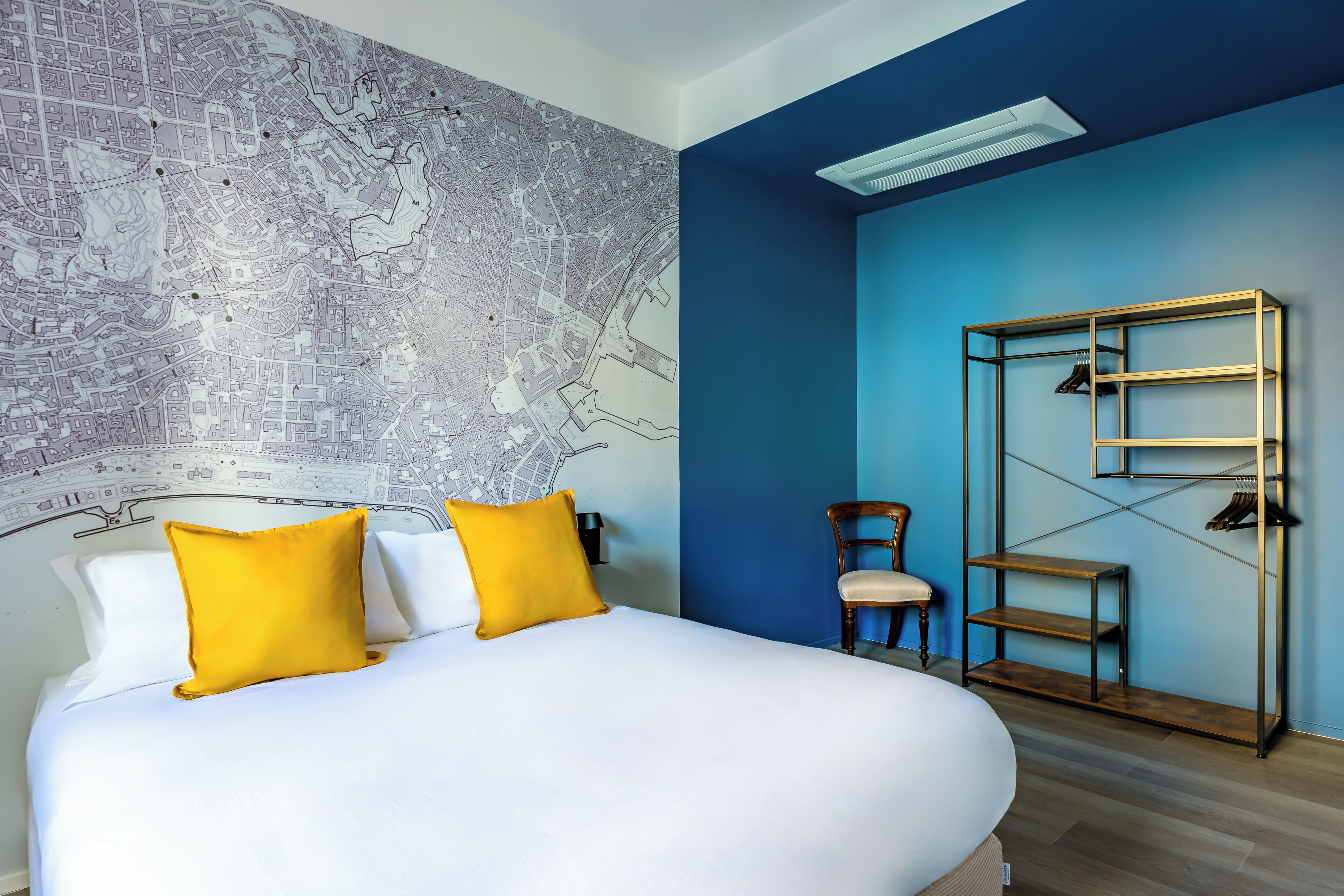 A hotel room with a large bed, two yellow pillows, a map mural on the wall, a blue accent wall, a wooden chair, and a metal shelf unit.