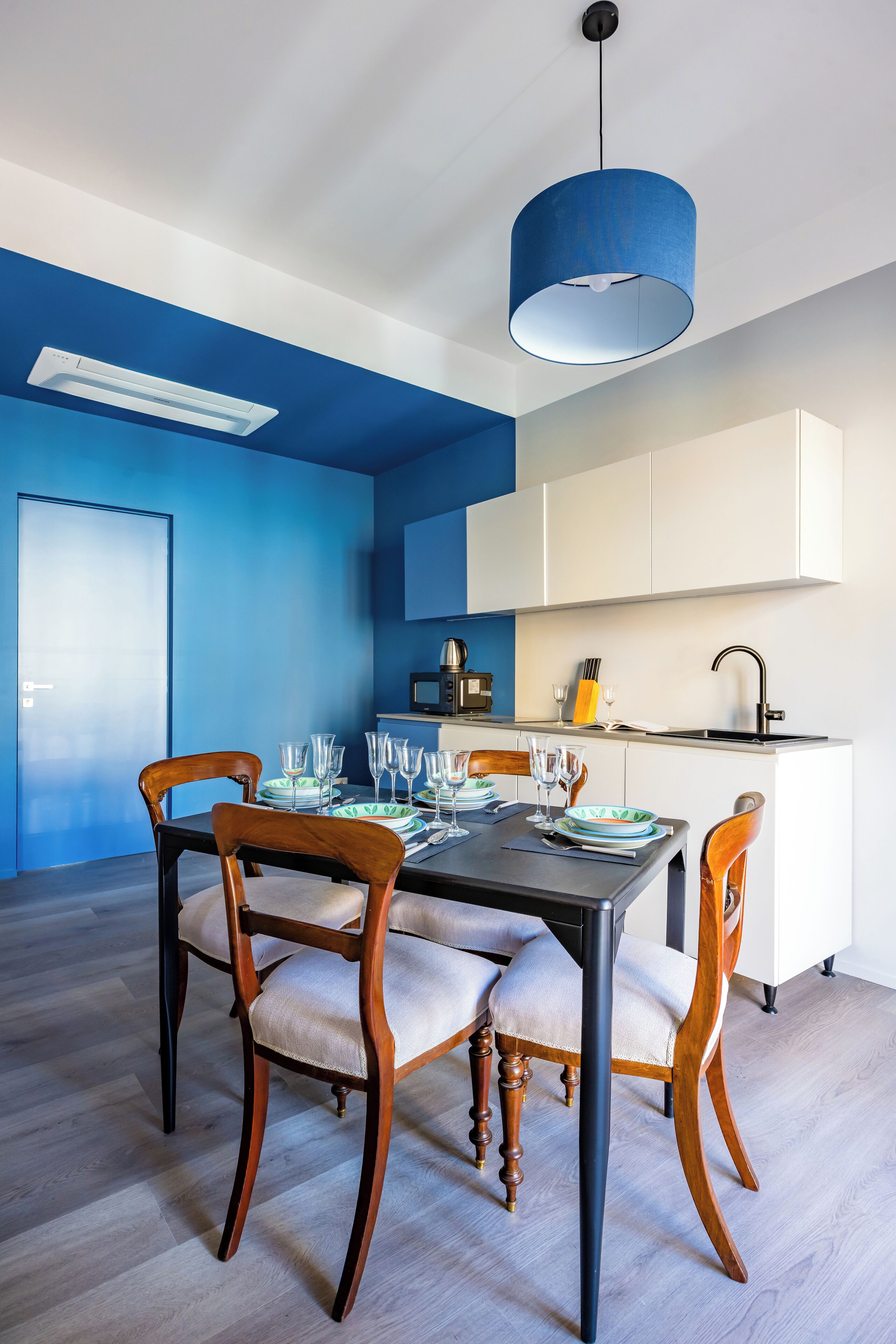 A dining area with a table set for four, wooden chairs, a blue pendant light, and a white cabinet in the background.