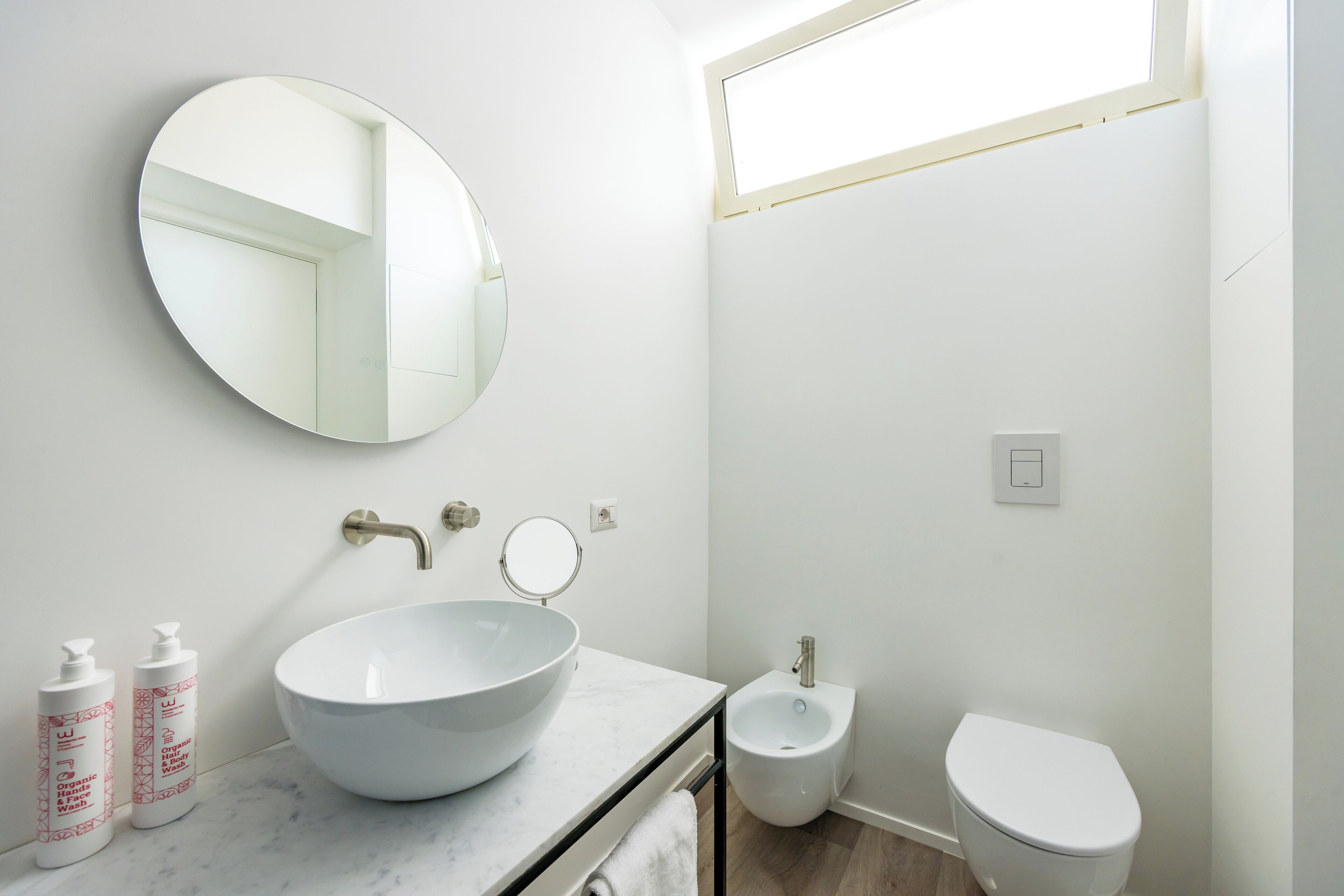 Ein Badezimmer mit rundem Spiegel, Waschbecken in Weiß, einem Bidet und zwei Shampoo-Flaschen.
