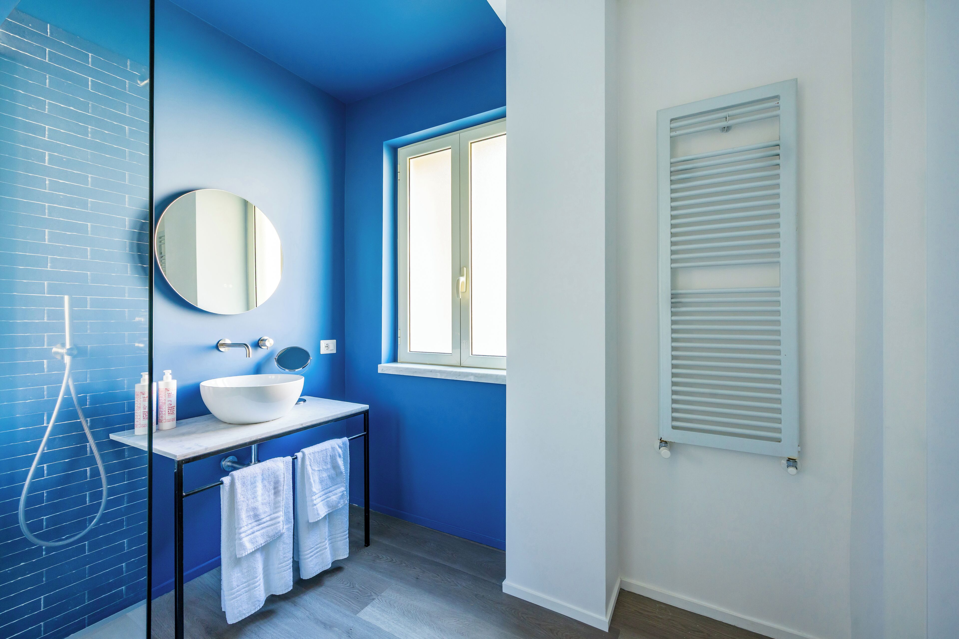 Une salle de bain avec un mur bleu, un lavabo blanc, un miroir rond et une fenêtre avec des volets blancs.