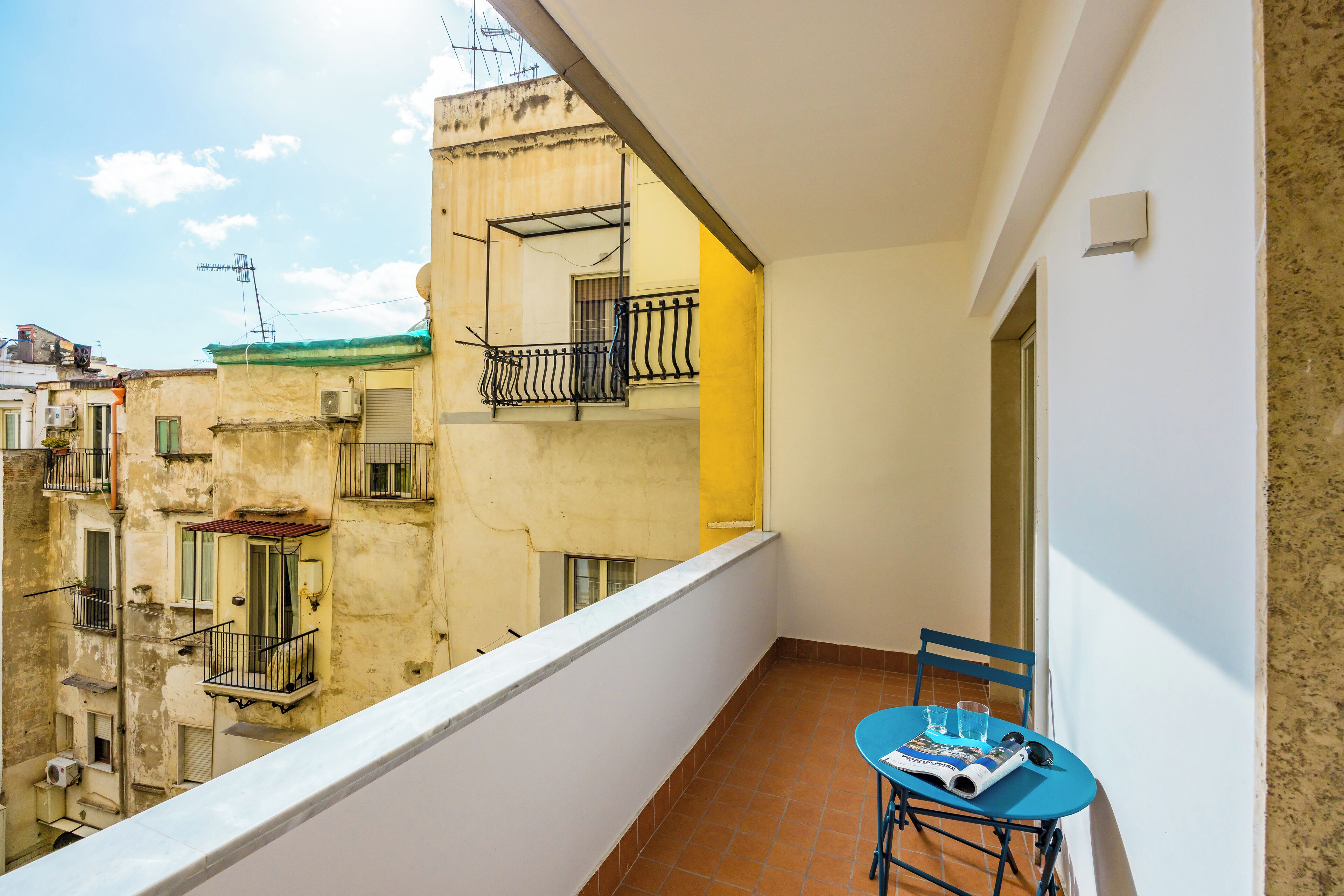 A balcony with a blue table and chair, a view of neighboring buildings, and a white wall.
