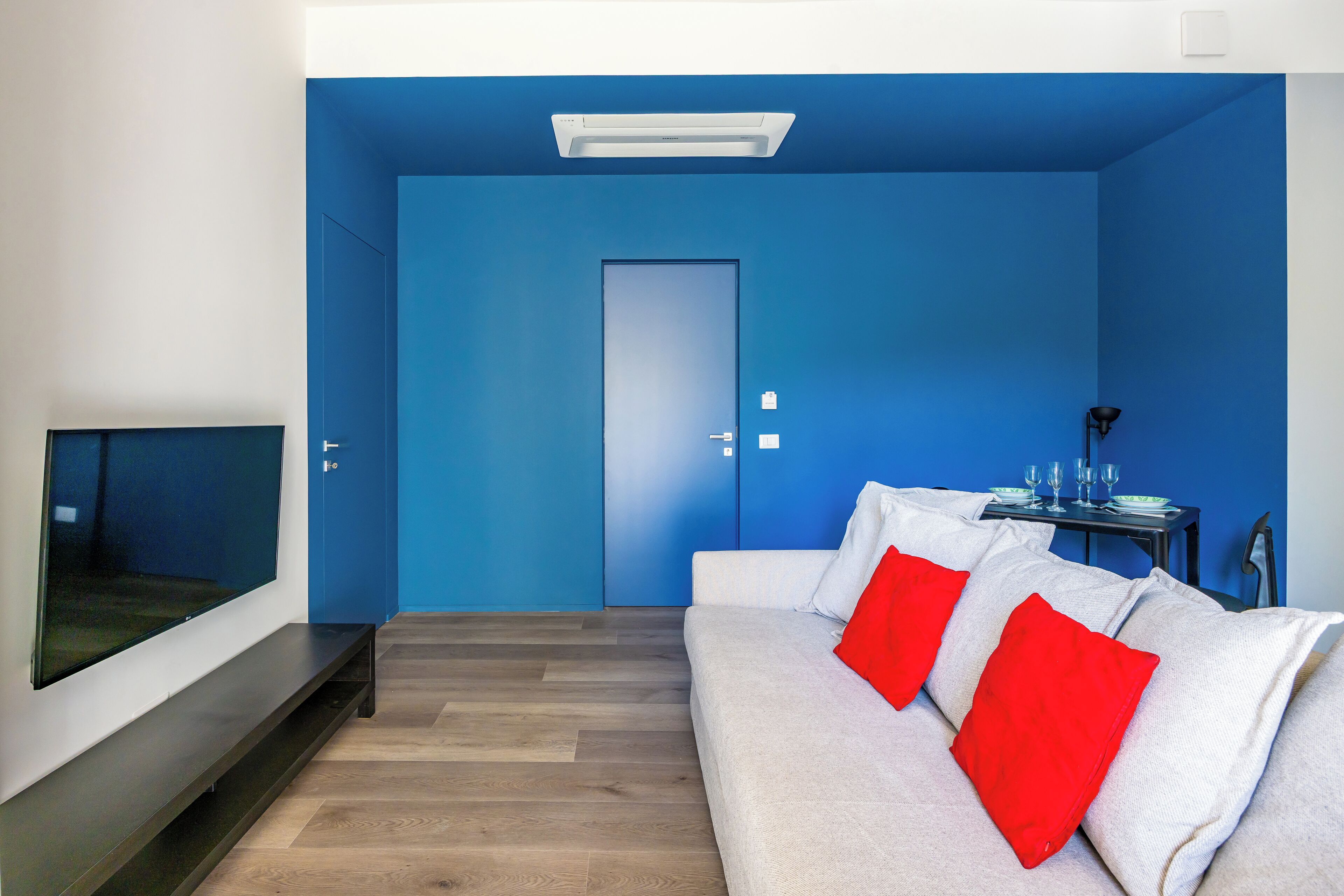 A modern living room with a blue accent wall, a grey sofa with red cushions, a flat-screen TV, and a small table with glasses and a green plate.