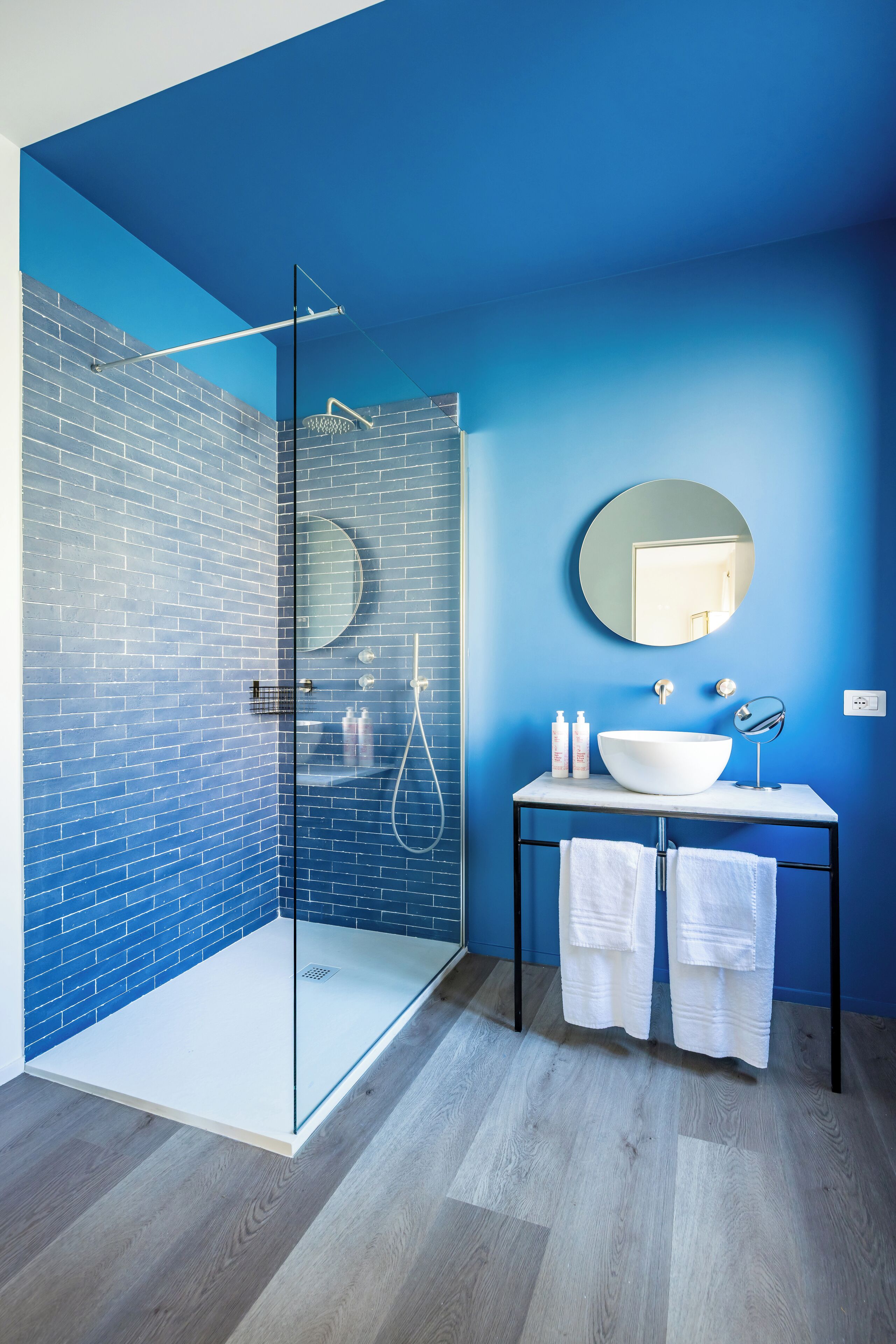 A bathroom with a glass-enclosed shower, a round mirror, a white sink, and a blue-tiled wall.