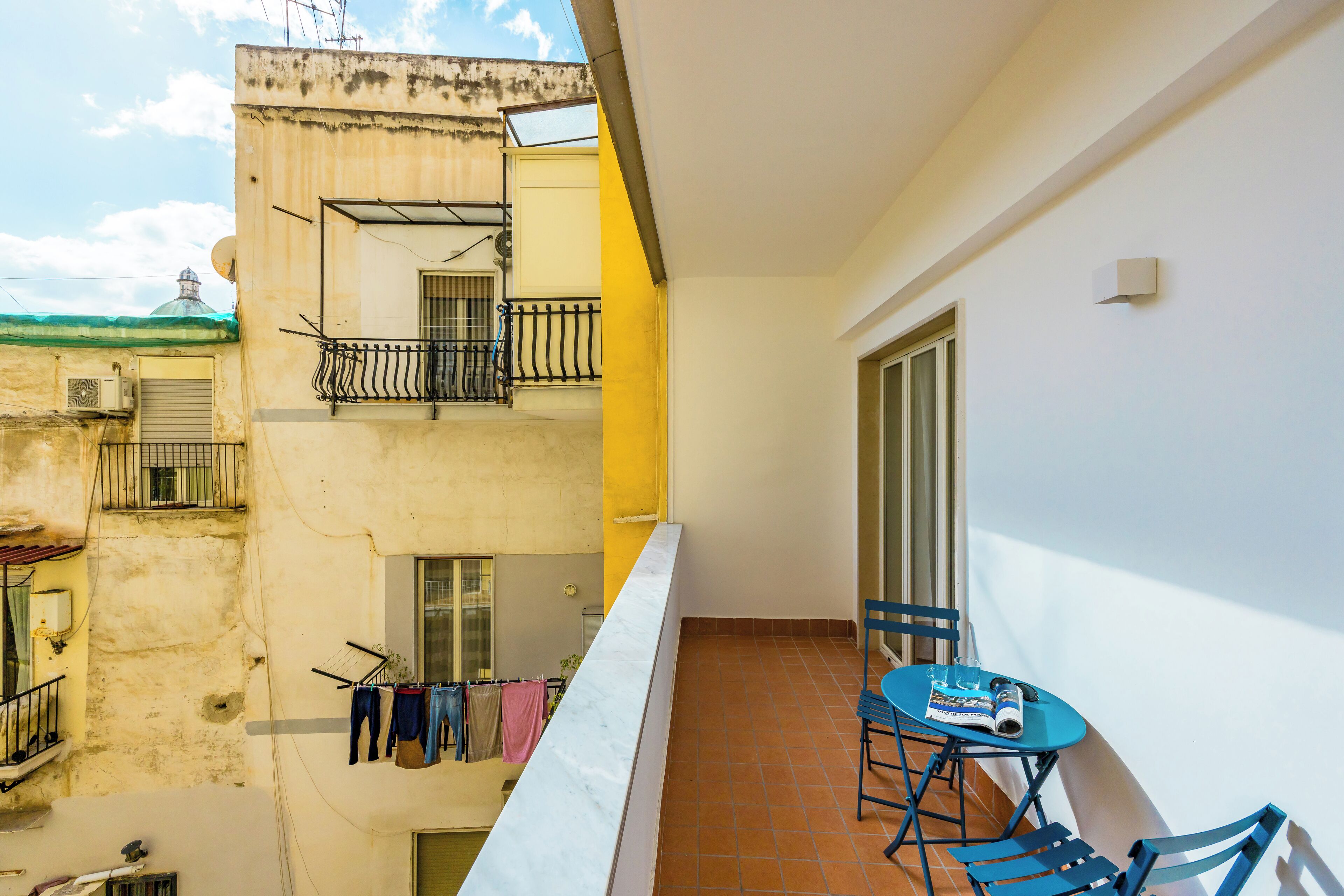 A balcony with a table and chairs, a clothesline with hanging clothes, and a view of neighboring buildings.