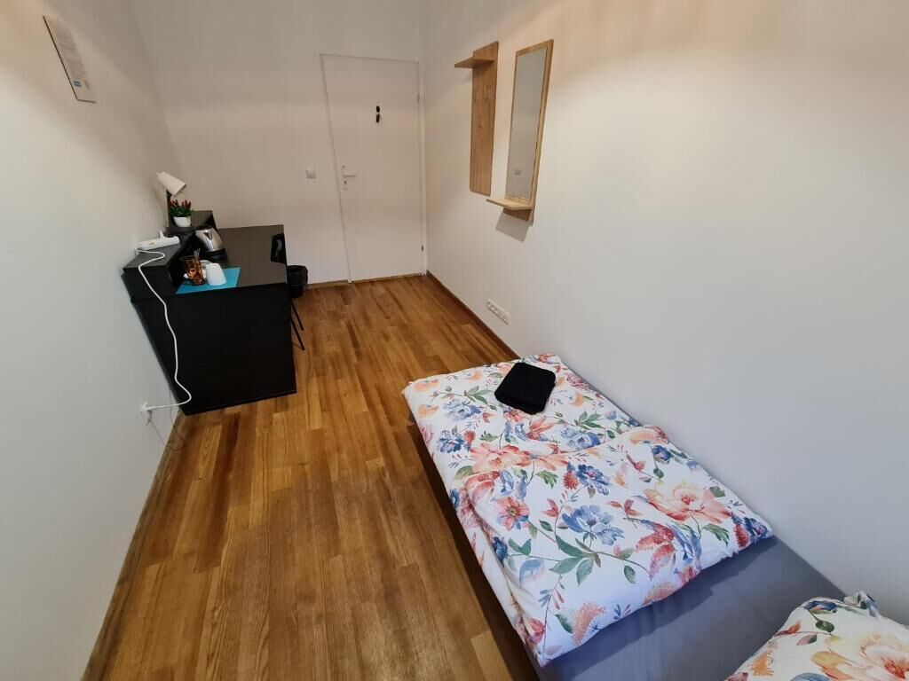A small, sparsely furnished room with a wooden floor, a desk with a chair, a bed with a floral bedspread, and a wall-mounted mirror.
