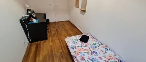A small, sparsely furnished room with a wooden floor, a desk with a chair, a bed with a floral bedspread, and a wall-mounted mirror.