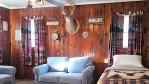 A room with wooden walls, a mounted deer head, plaid curtains, a sofa, a bed with a patterned blanket, and a sign that reads "MAN CAVE". - The Huntsmen (Hillsboro)
