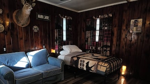 A room with a wooden floor, a blue sofa, a bed with a patterned quilt, and a deer head mounted on the wall. - The Huntsmen (Hillsboro)