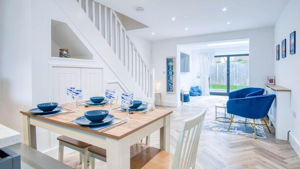 A modern dining area with a wooden table set for four, white chairs, and a staircase leading to an upper level. - Hatherleigh Drive - 2 Bedroom House - Mumbles (Swansea)